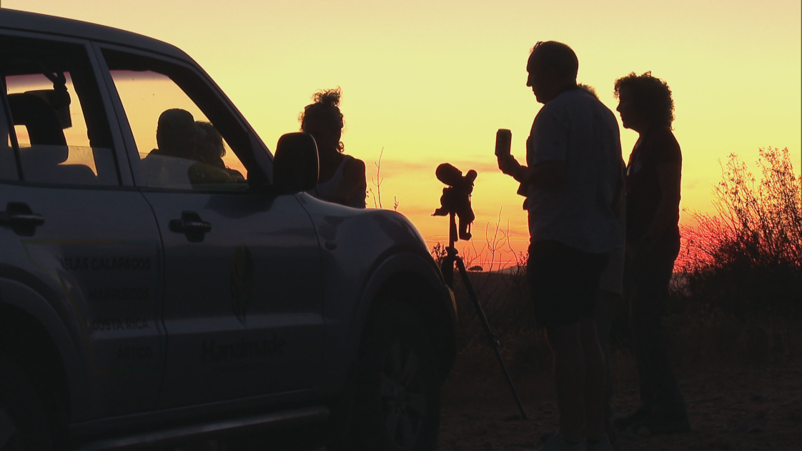 Siluetas de personas y un vehículo al atardecer, con un trípode y equipo de observación. Cielo anaranjado y rojizo. Naturaleza.
