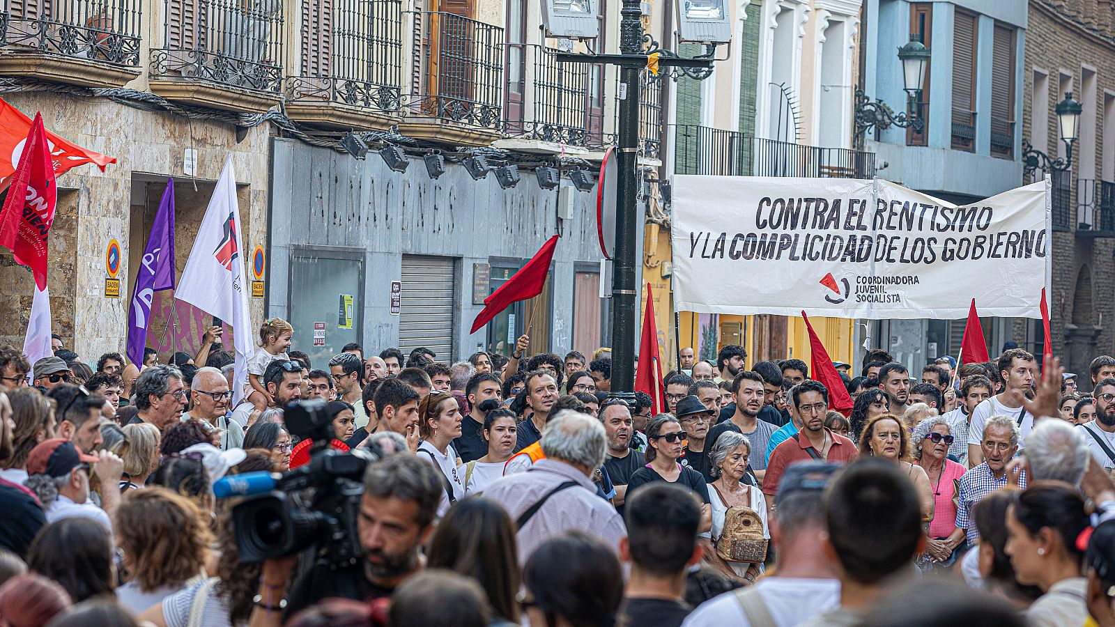 Manifestación urbana con multitud diversa, banner "CONTRA EL RENTISMO Y LA COMPLICIDAD DE LOS GOBIERNOS" y logo "COORDINADORA JUVENIL SOCIALISTA". Banderas rojas, blancas y moradas presentes. Persona con cámara de vídeo profesional.