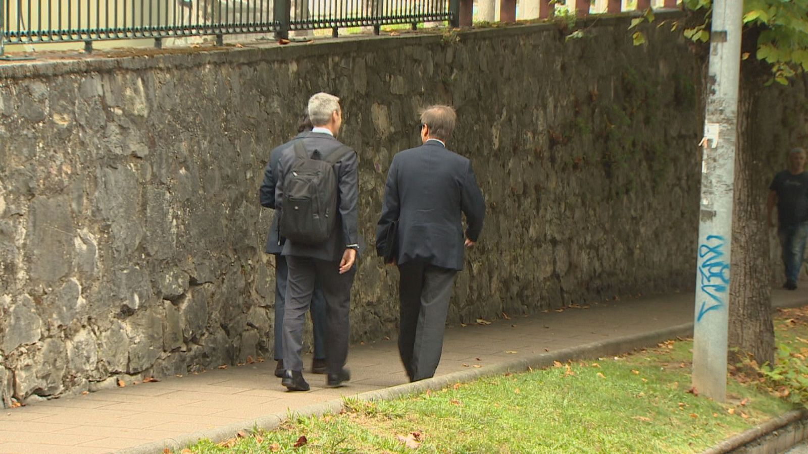 Dos hombres con trajes oscuros caminan por una acera. Uno tiene cabello canoso y mochila negra. A la derecha, un poste de luz con grafitis azules.