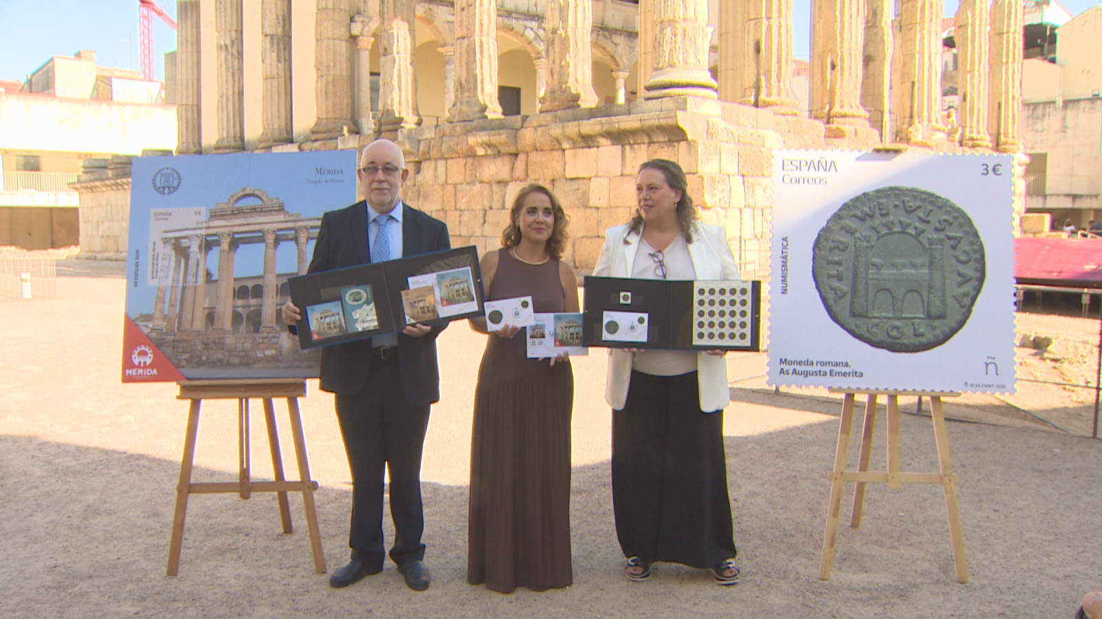 Presentación de sellos postales numismáticos en Mérida, España, con ruinas romanas de fondo. Tres personas exhiben colecciones filatélicas y numismáticas.