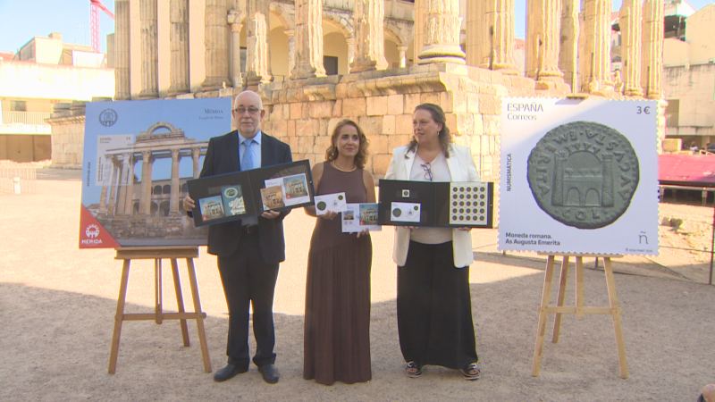 Presentación de sellos postales numismáticos en Mérida, España, con ruinas romanas de fondo. Tres personas exhiben colecciones filatélicas y numismáticas.