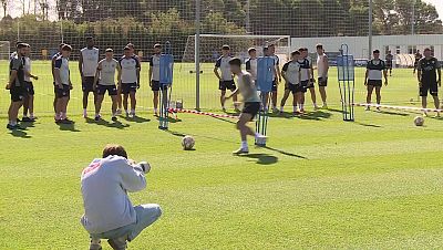 Entrenamiento de f�tbol con fot�grafo en primer plano, jugador practicando golpeo de bal�n y equipo observando al fondo. C�sped verde y cielo despejado.