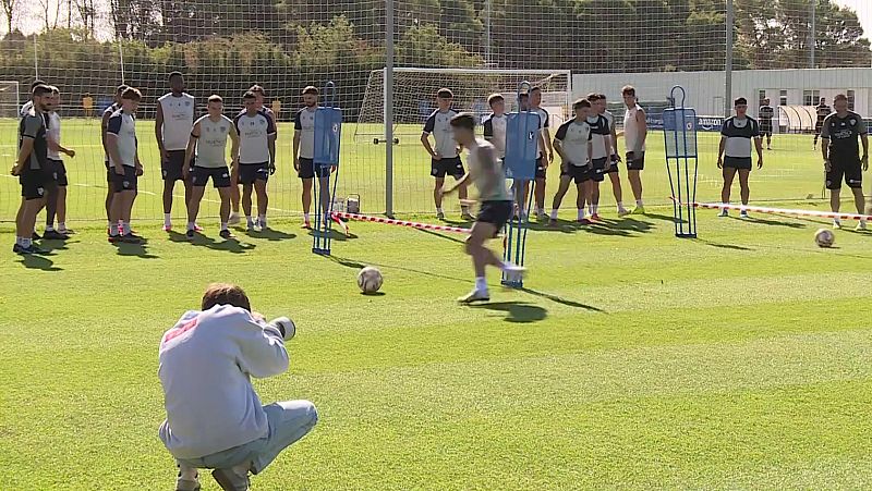 Entrenamiento de fútbol con fotógrafo en primer plano, jugador practicando golpeo de balón y equipo observando al fondo. Césped verde y cielo despejado.