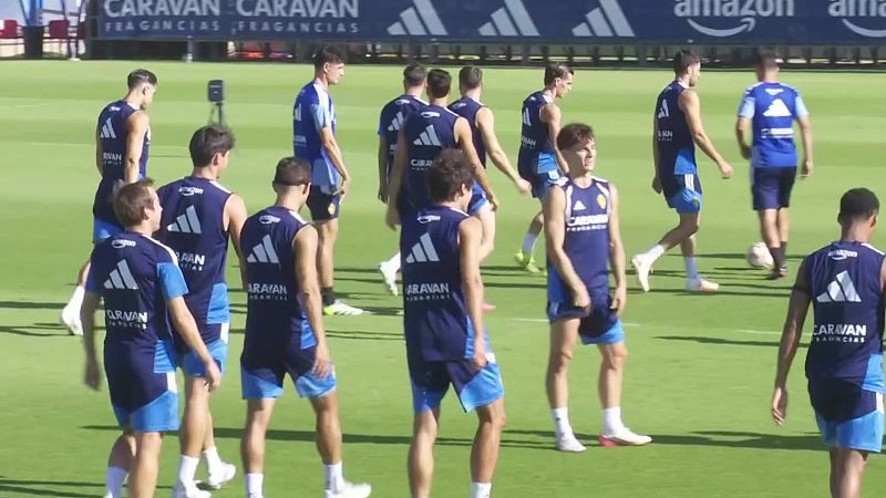 Jugadores atléticos con uniformes azul oscuro y blanco, luciendo logos de Adidas y patrocinadores como "CARAVAN FRAGANCIAS" y "AMAZON", entrenan en un campo de césped verde.