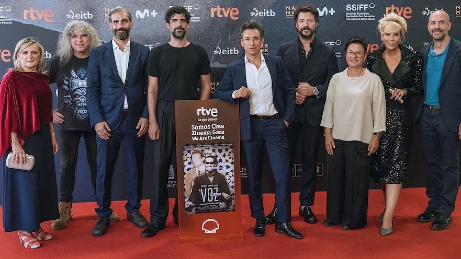 En el Festival de San Sebastián, Jon Sistiaga, David Trueba, Roberto Leal, Álvaro Morte, Ana Pardo de Vera, Alaska, Antonio de la Torre, Mario Vaquerizo y una mujer posan junto al standee de "LA VOZ QUEBRADA".