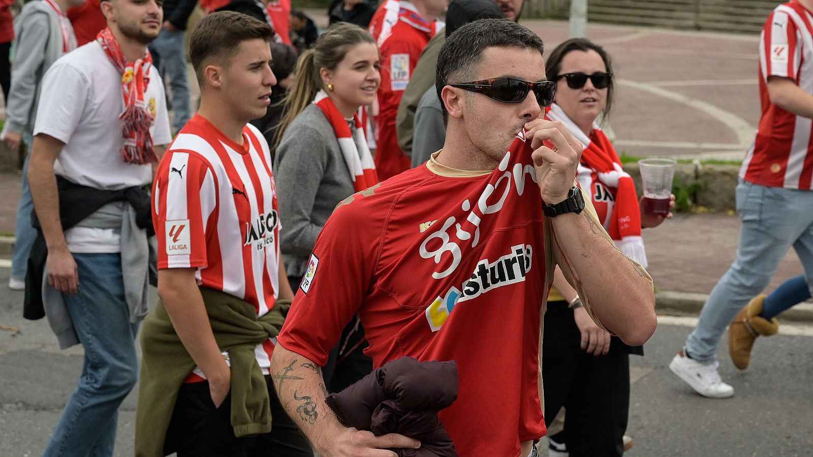 Aficionados del Real Sporting de Gijón, con camisetas rojas y blancas, apoyan al equipo en la calle. Destaca un hombre con "Gijón Asturias" en su camiseta, animando.