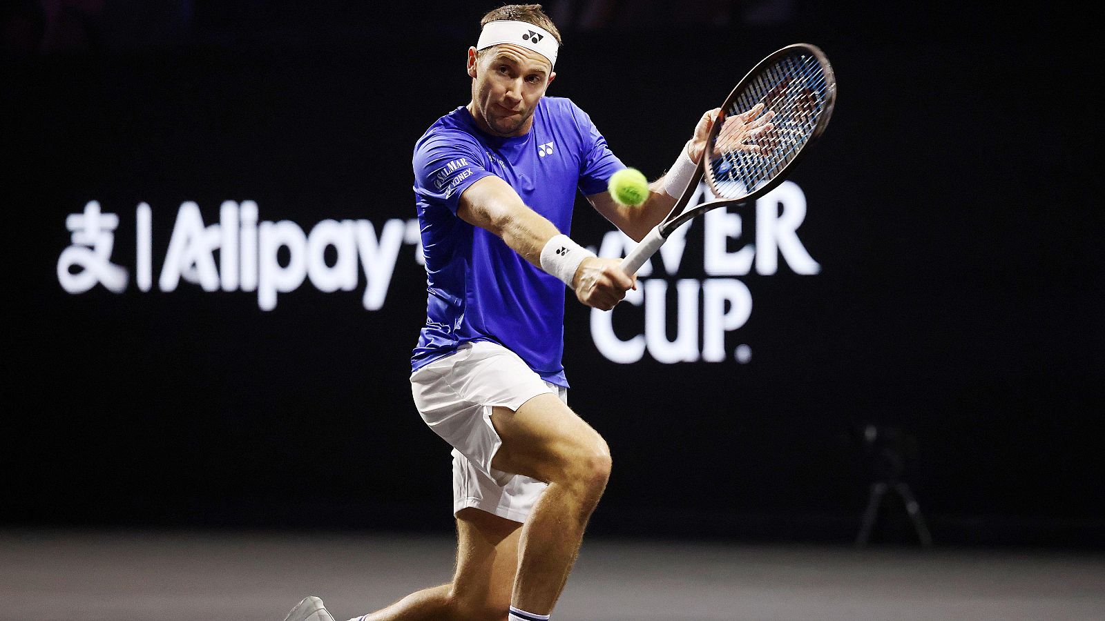 Casper Ruud golpea una pelota de tenis con su raqueta, concentrado. Viste camiseta azul, pantalón blanco y cinta Yonex. Al fondo, logotipos de Alipay y Laver Cup.