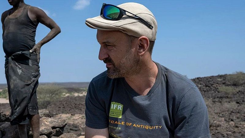 Hombre con gorra y camiseta del "INSTITUTE FOR FIELD RESEARCH" en un paisaje rocoso. A su lado, otra persona con vestimenta oscura.