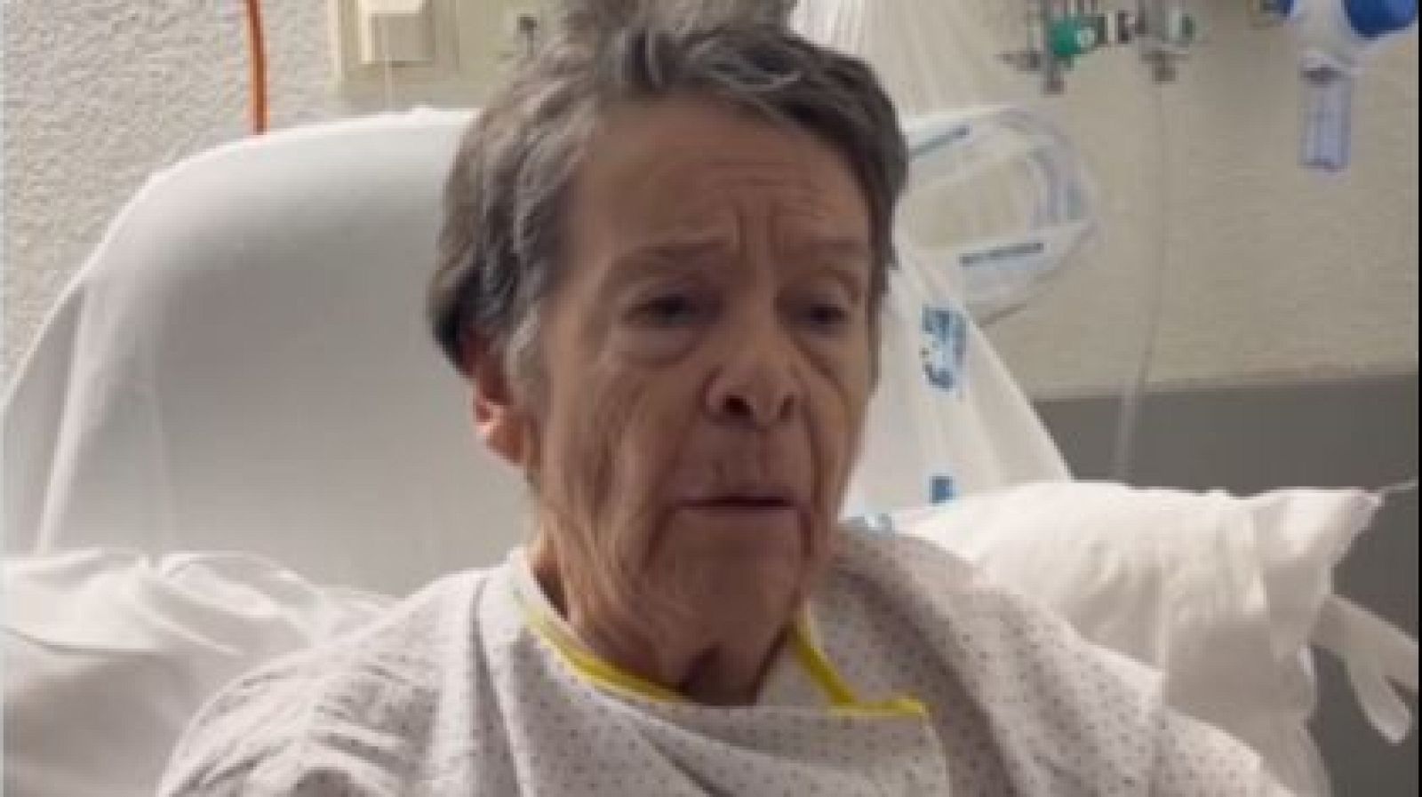Mari Carmen, activista de la PAH, en una habitación de hospital con bata blanca y azul, hace un llamamiento a la movilización por la vivienda. Se aprecian equipos médicos al fondo.