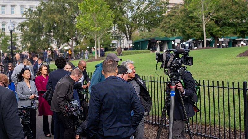 Periodistas y equipo de prensa se congregan en los exteriores de la Casa Blanca, con un operador de cámara listo y una valla separando al grupo del edificio.