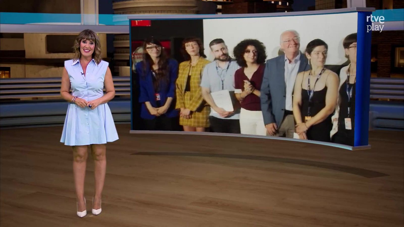 Mónica López en estudio de RTVE con vestido azul claro. Pantalla digital muestra grupo de personas con lanyards en contexto de "Loterías".