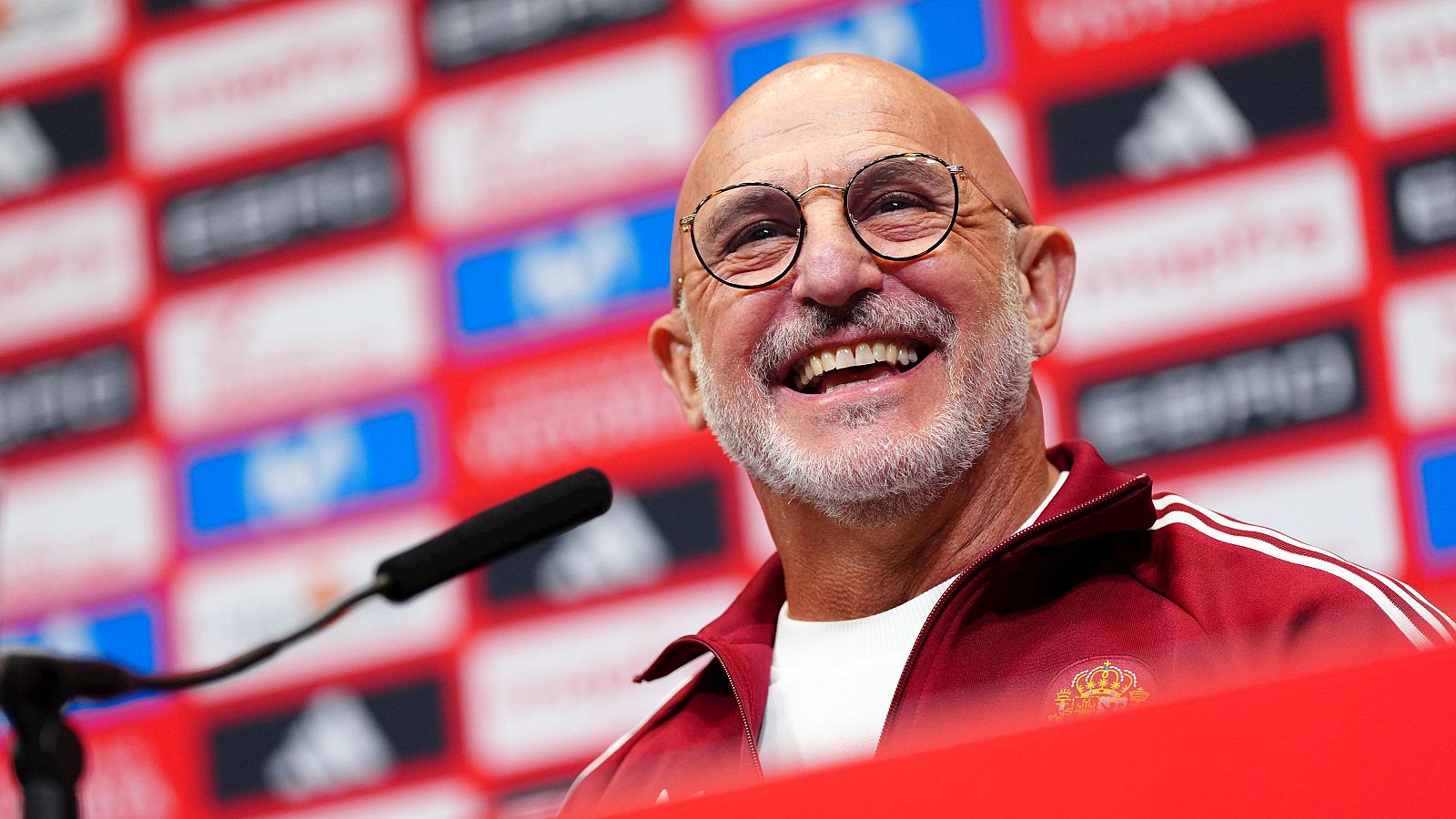 Luis de la Fuente, seleccionador español, sonríe en rueda de prensa con chaqueta granate y escudo RFEF. Fondo rojo con logos.