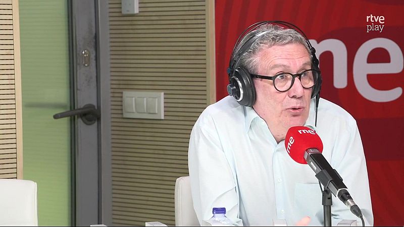 Luisgé Martín en un estudio de radio de RNE, con micrófono rojo y logo de RTVE Play al fondo. Lleva gafas, auriculares y camisa clara, y está hablando.