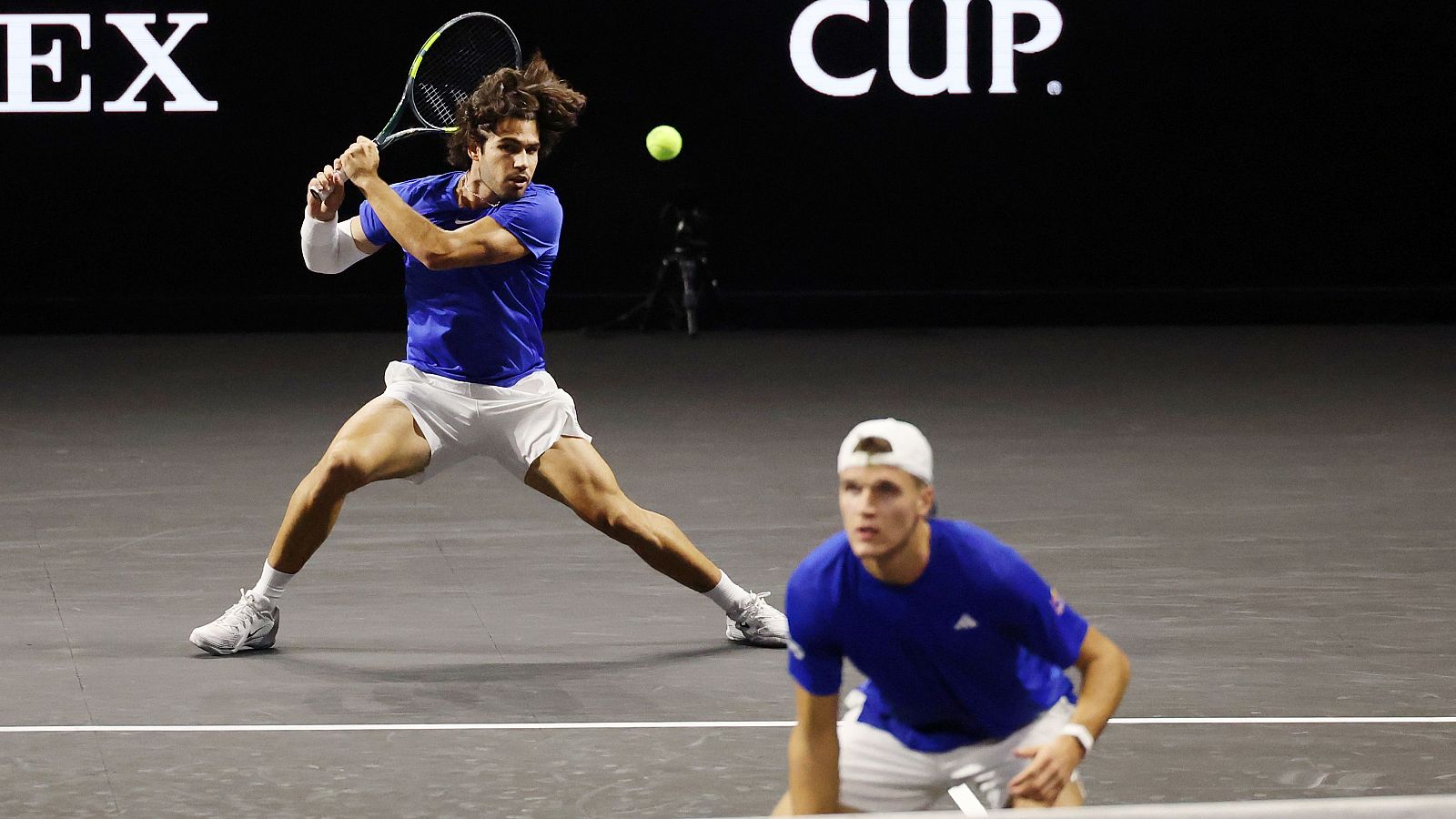 Carlos Alcaraz golpea un revés a dos manos en una cancha de tenis oscura. Se observa a otro jugador en segundo plano y logos de patrocinadores como ROLEX y CUP.
