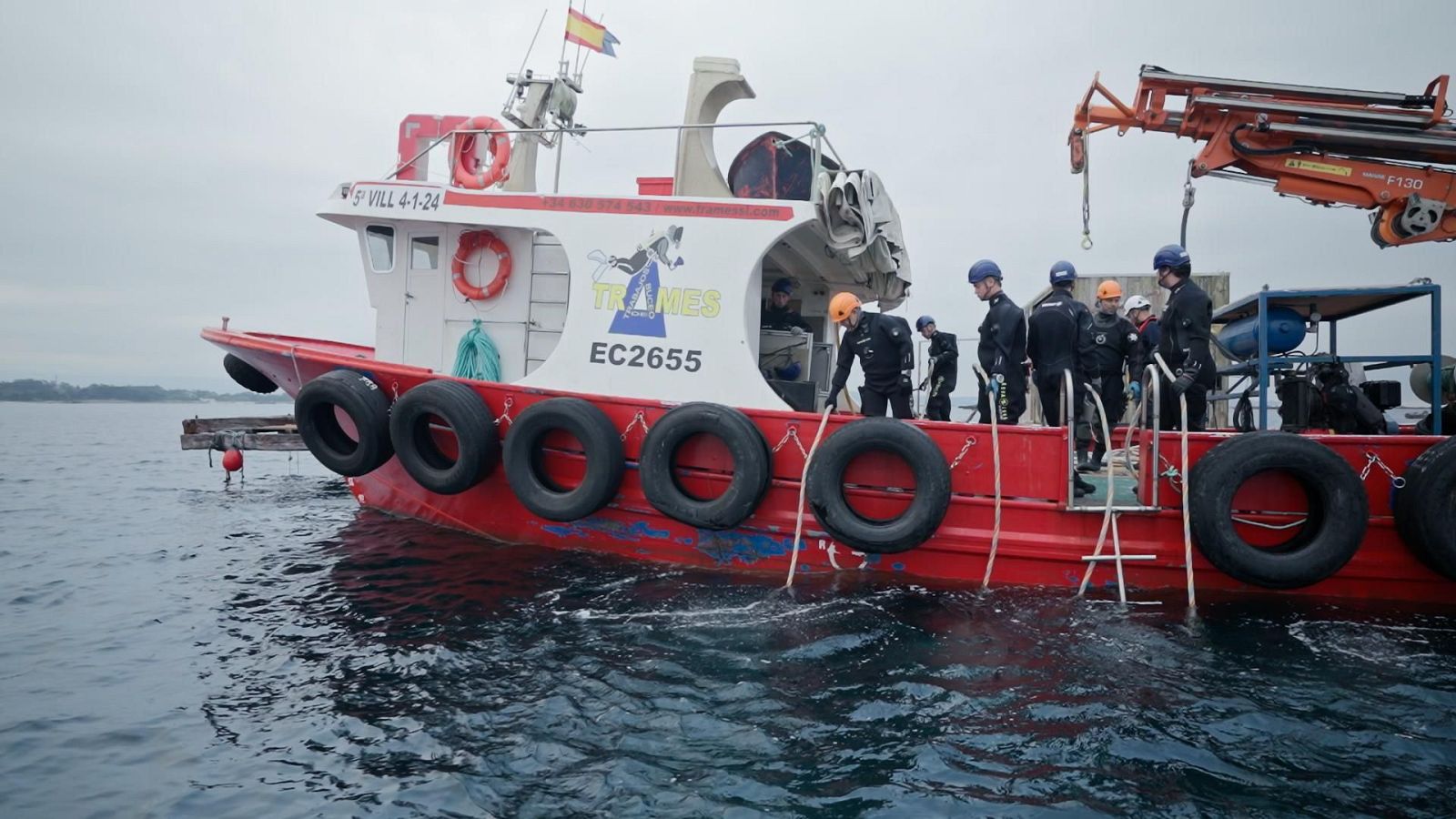 Embarcación de trabajo roja y blanca con personal uniformado y cascos, operando en aguas tranquilas bajo cielo nublado. Se aprecian inscripciones "S VILL 4-1-24", "TRITONES EC2655" y grúa "F130".