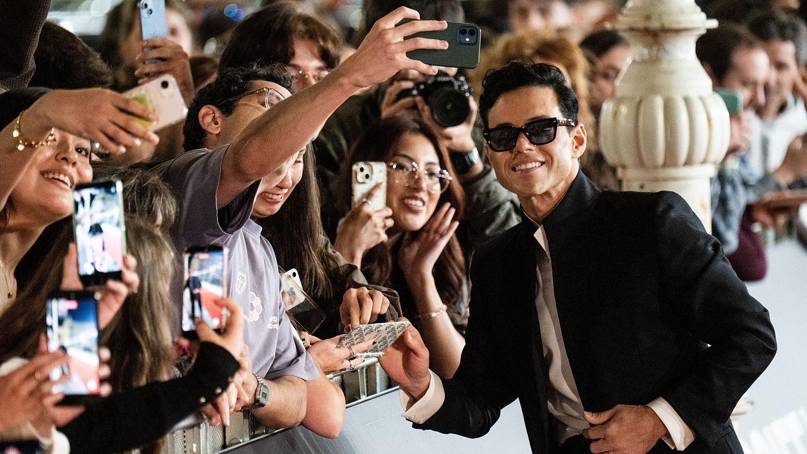 Rami Malek interactúa con fans en un evento, sonriendo y saludando. La multitud, emocionada, toma fotos con sus móviles. Se observa una barrera y un pilar blanco al fondo.