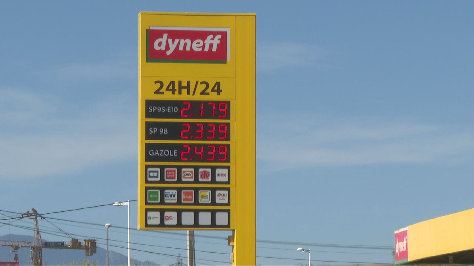 Cartel de gasolinera Dyneff con precios de combustible (SP95-E10: 2.179, SP98: 2.339, Gazole: 2.439) y logotipos de pago bajo cielo azul.