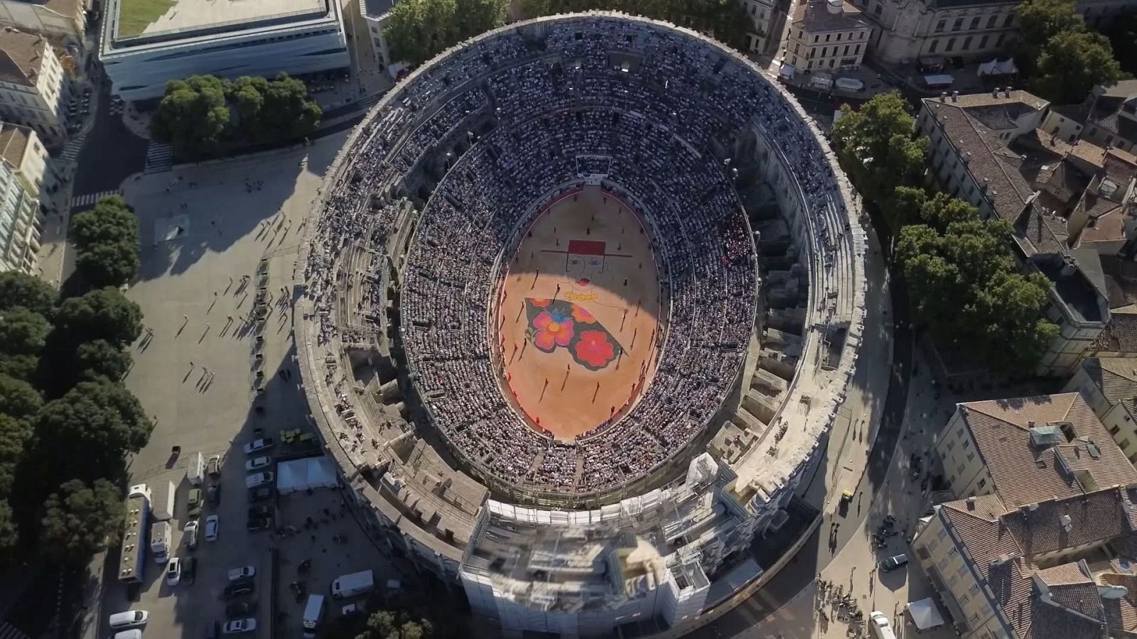 Plaza de toros ovalada con público en gradas y un gran diseño colorido en el ruedo, rodeada de ciudad. Asociada a "Tendido Cero", PGM 429, 26 de septiembre.