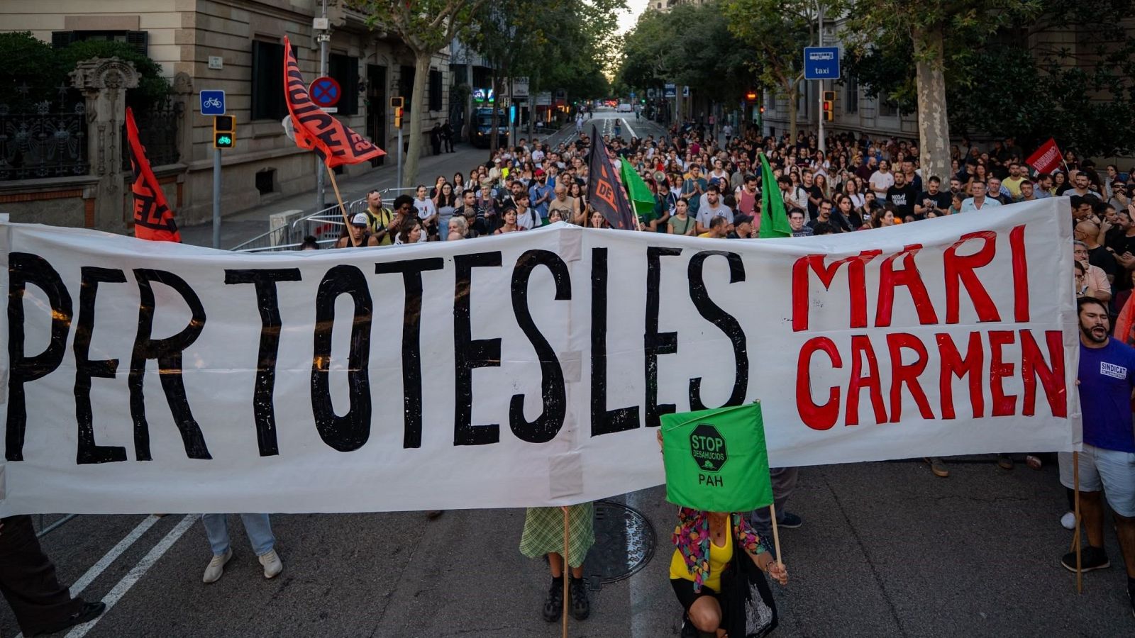 Manifestación urbana con pancarta "PER TOTES LES MARI CARMEN" y bandera "STOP DESAHUCIOS PAH", protestando contra desahucios.