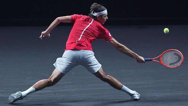 Cameron Norrie, con pelo largo recogido y cinta blanca, golpea una raqueta naranja rojiza en un movimiento de derecha potente. Viste camiseta roja y pantal�n gris. Pelota amarilla en el aire.