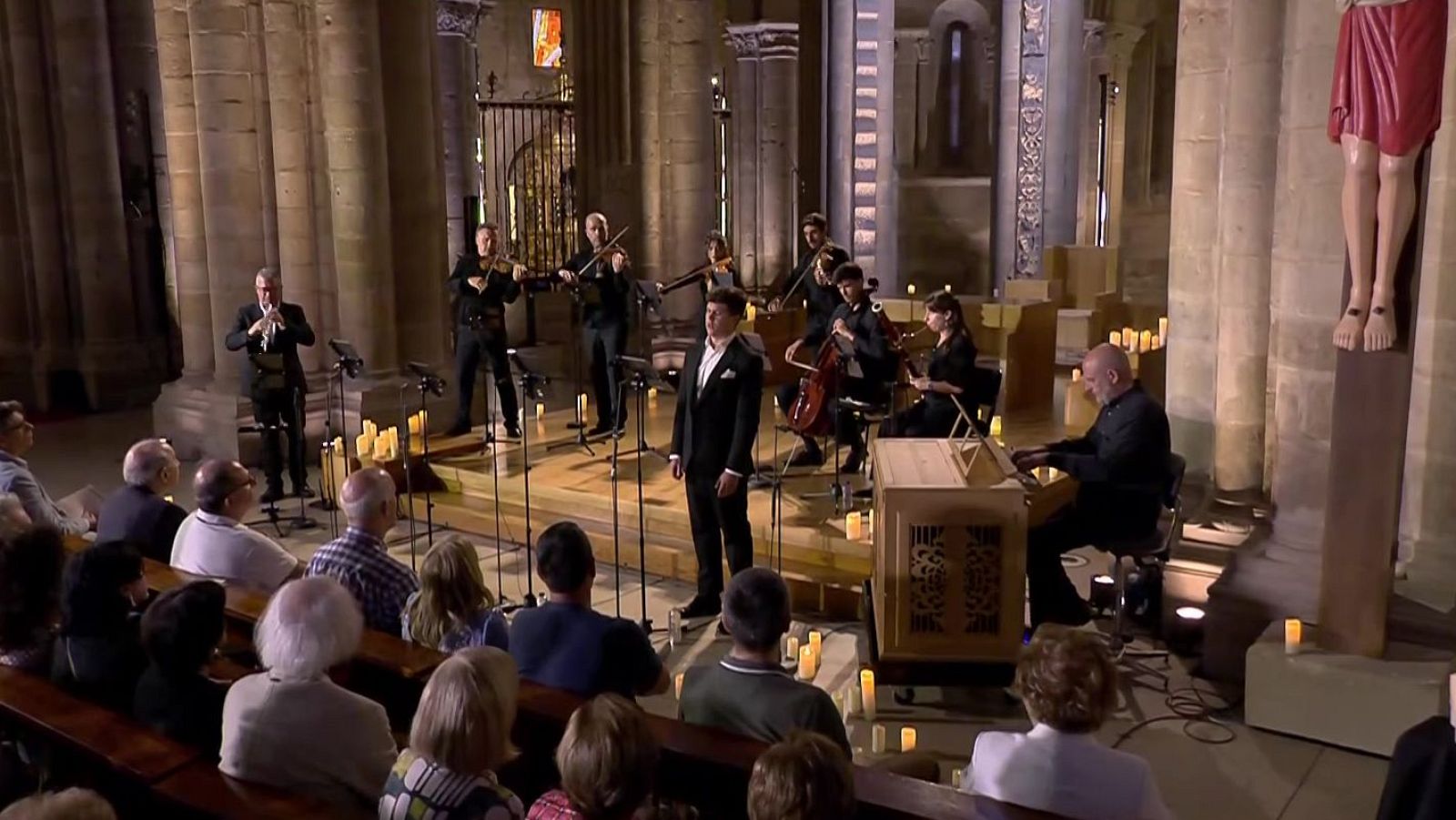 Concierto de música clásica en una iglesia antigua con cantante, flautista, violinistas, violonchelista, clavecinista y organista, ante un público atento.