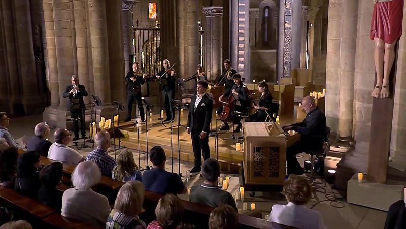 Concierto de música clásica en una iglesia antigua con cantante, flautista, violinistas, violonchelista, clavecinista y organista, ante un público atento.