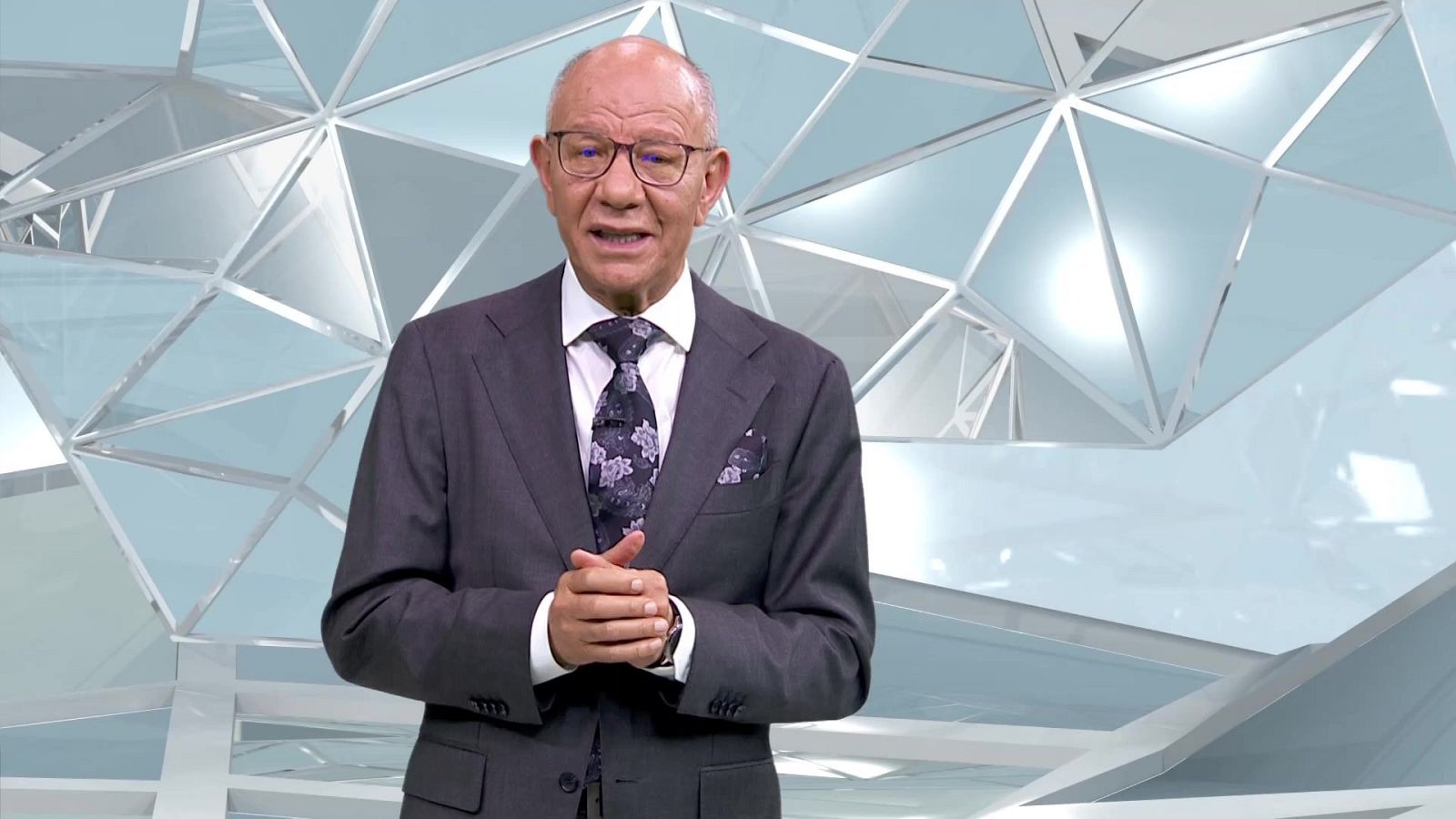 Fernando Medina, presentador de TVE, posa con traje gris, camisa blanca y corbata floral en un set de televisión con fondo geométrico azul y blanco.