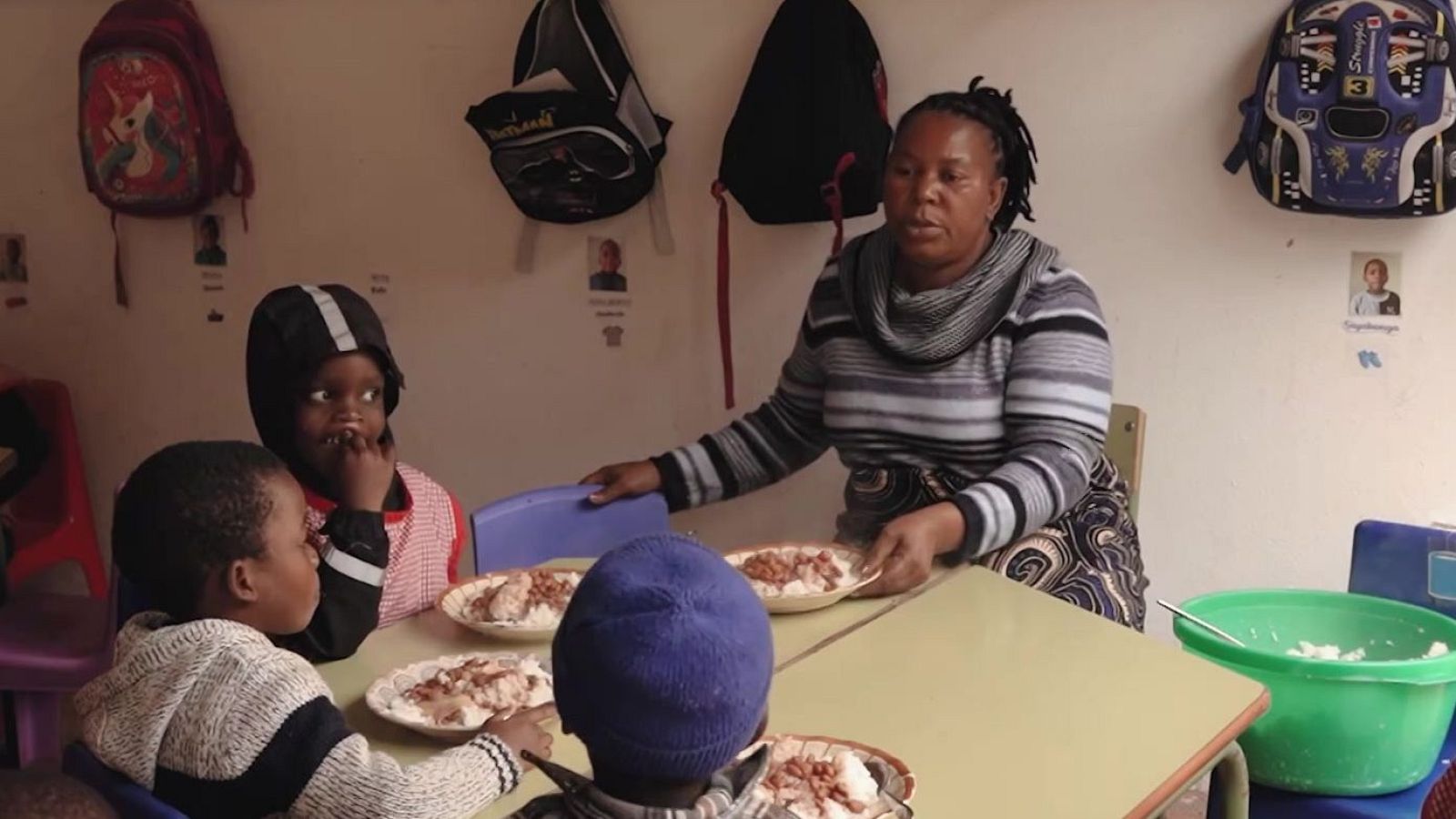 Mujer adulta sirve comida a niños en un comedor infantil. Se ven platos con arroz y guiso, mochilas y dibujos en la pared. Entorno dedicado a los niños.