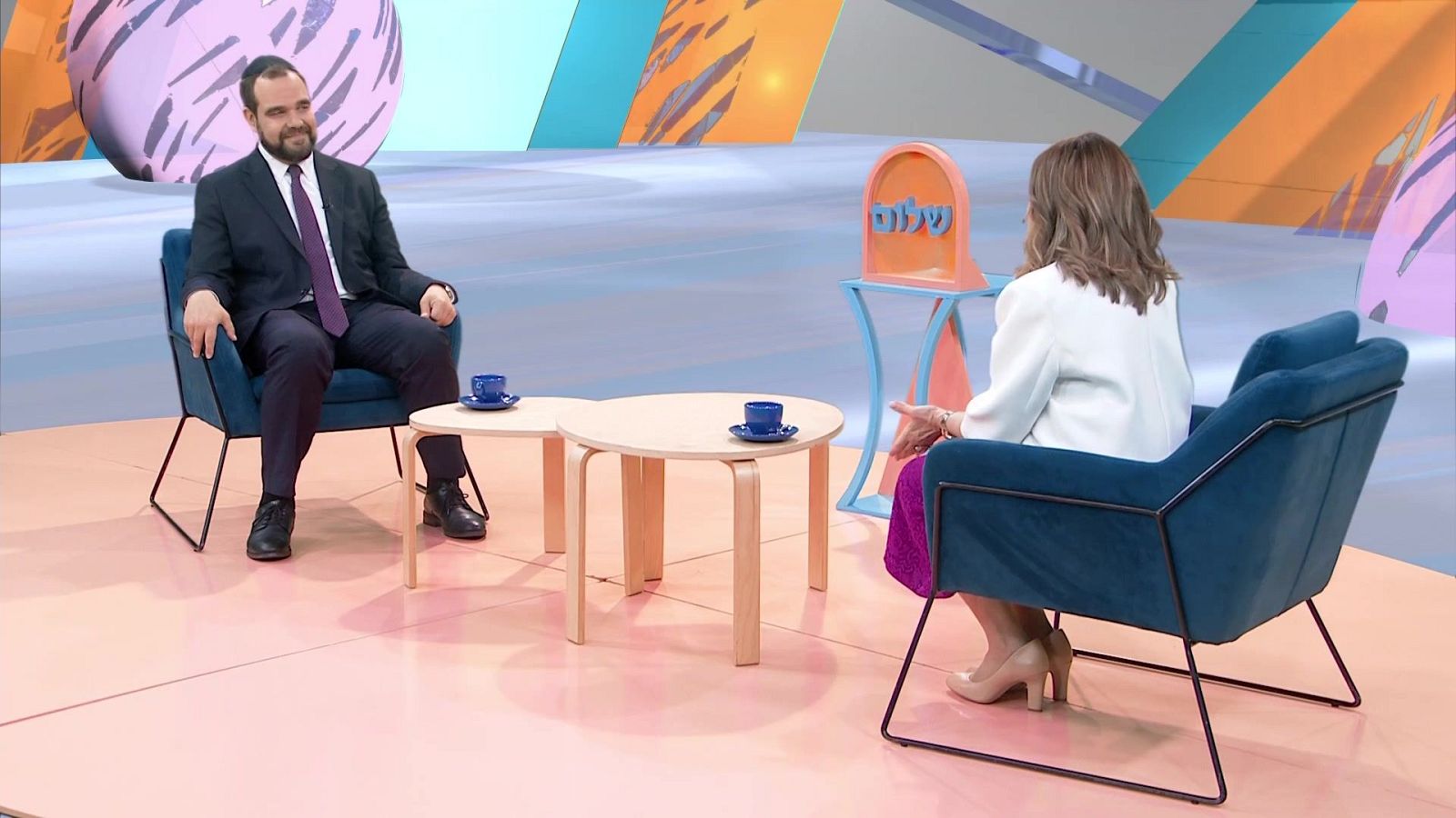 Jon Sistiaga conversa con una mujer en un estudio de televisión. Detrás de ella, un objeto decorativo muestra la palabra "שלום" en hebreo.