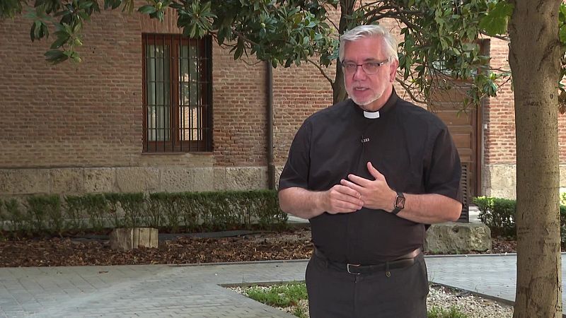 Sacerdote con alzacuellos y gafas, de pie frente a un edificio antiguo de ladrillo en un jardín. Transmite solemnidad.