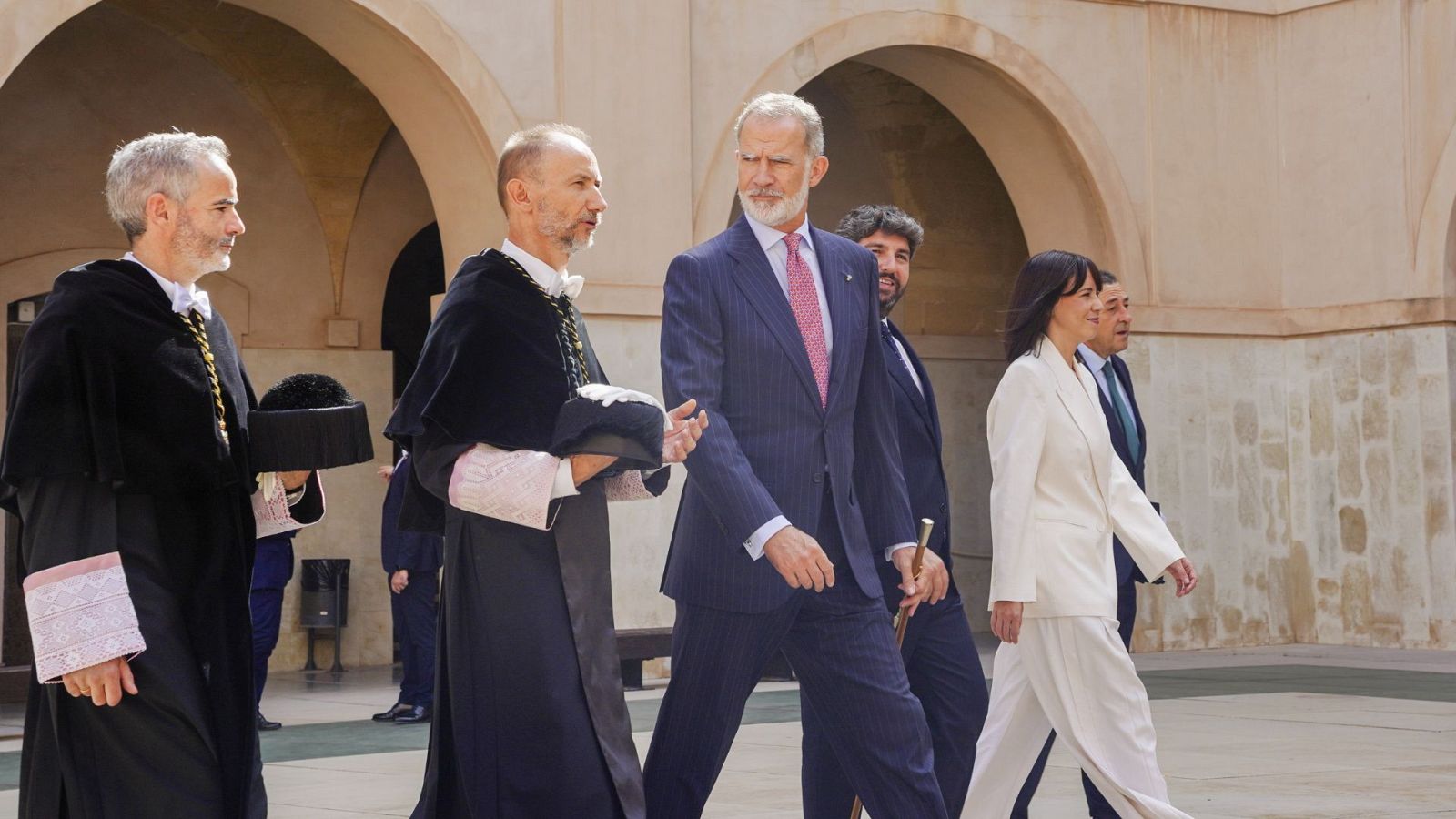 Felipe VI, Rey de España, camina por un patio histórico con arcos de piedra, acompañado por Pilar Llop y dos dignatarios académicos. El Rey viste traje azul oscuro y Llop, traje blanco roto.