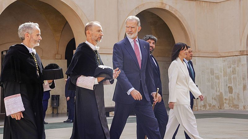 Felipe VI, Rey de España, camina por un patio histórico con arcos de piedra, acompañado por Pilar Llop y dos dignatarios académicos. El Rey viste traje azul oscuro y Llop, traje blanco roto.
