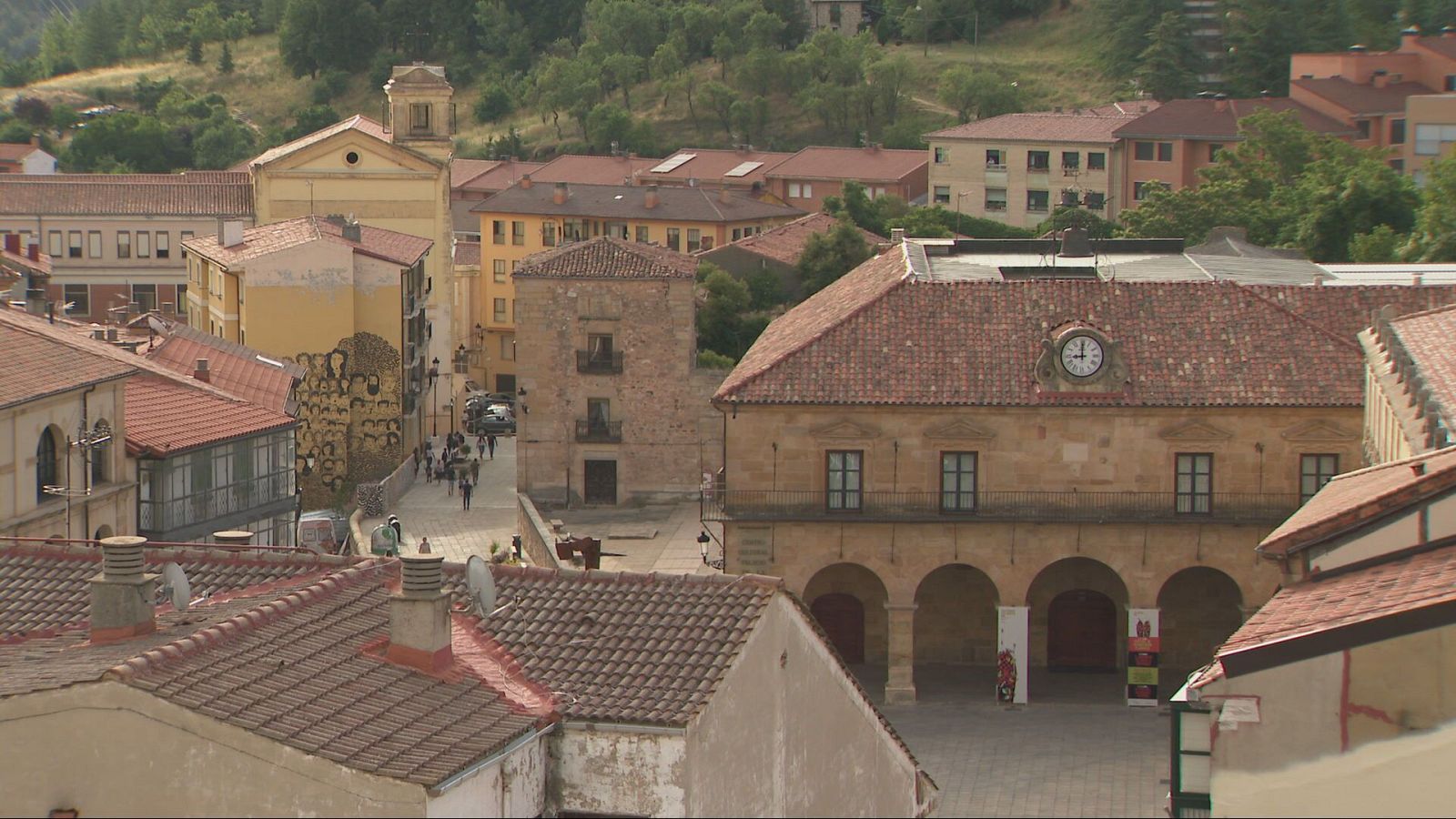 Vista panorámica de un pueblo español con arquitectura tradicional, arcos, un mural artístico y colinas verdes al fondo.