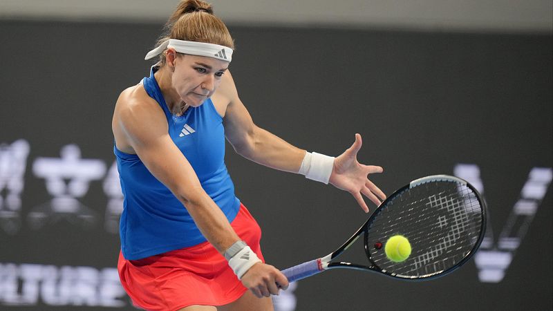 Aryna Sabalenka golpea una pelota de tenis amarilla con su raqueta, vistiendo indumentaria de Adidas: camiseta azul, falda roja y mu�equeras. Fondo oscuro con caracteres asi�ticos borrosos.