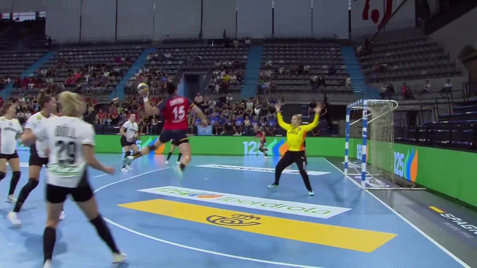 Jugadora de balonmano con dorsal 15 en rojo salta para lanzar. Portera de amarillo se prepara. En primer plano, jugadora con dorsal 29 y "ADUL".