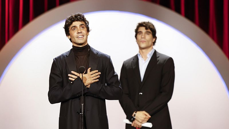Javier Ambrossi y Javier Calvo, directores españoles, en un escenario. Ambrossi habla a un micrófono con gesto emotivo, mientras Calvo sostiene un documento.