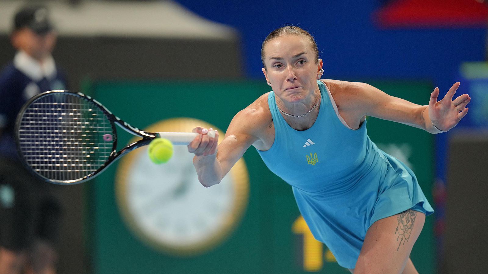 Elina Svitolina en acción, golpeando una pelota de tenis con raqueta. Viste vestido azul claro con Adidas y emblema de Ucrania. Tatuaje visible en muslo derecho.
