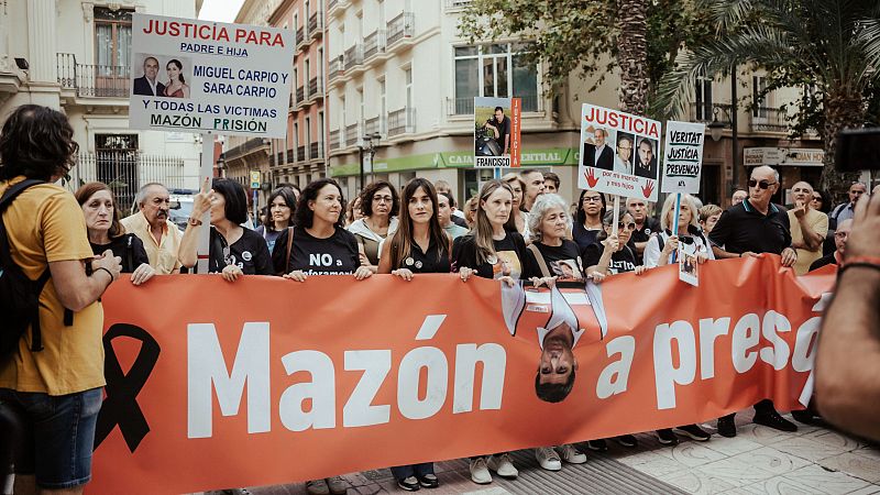 Manifestación con pancarta "Mazón a prisión" y foto invertida del presidente. Carteles de justicia y víctimas. Edificios urbanos y letrero bancario al fondo.