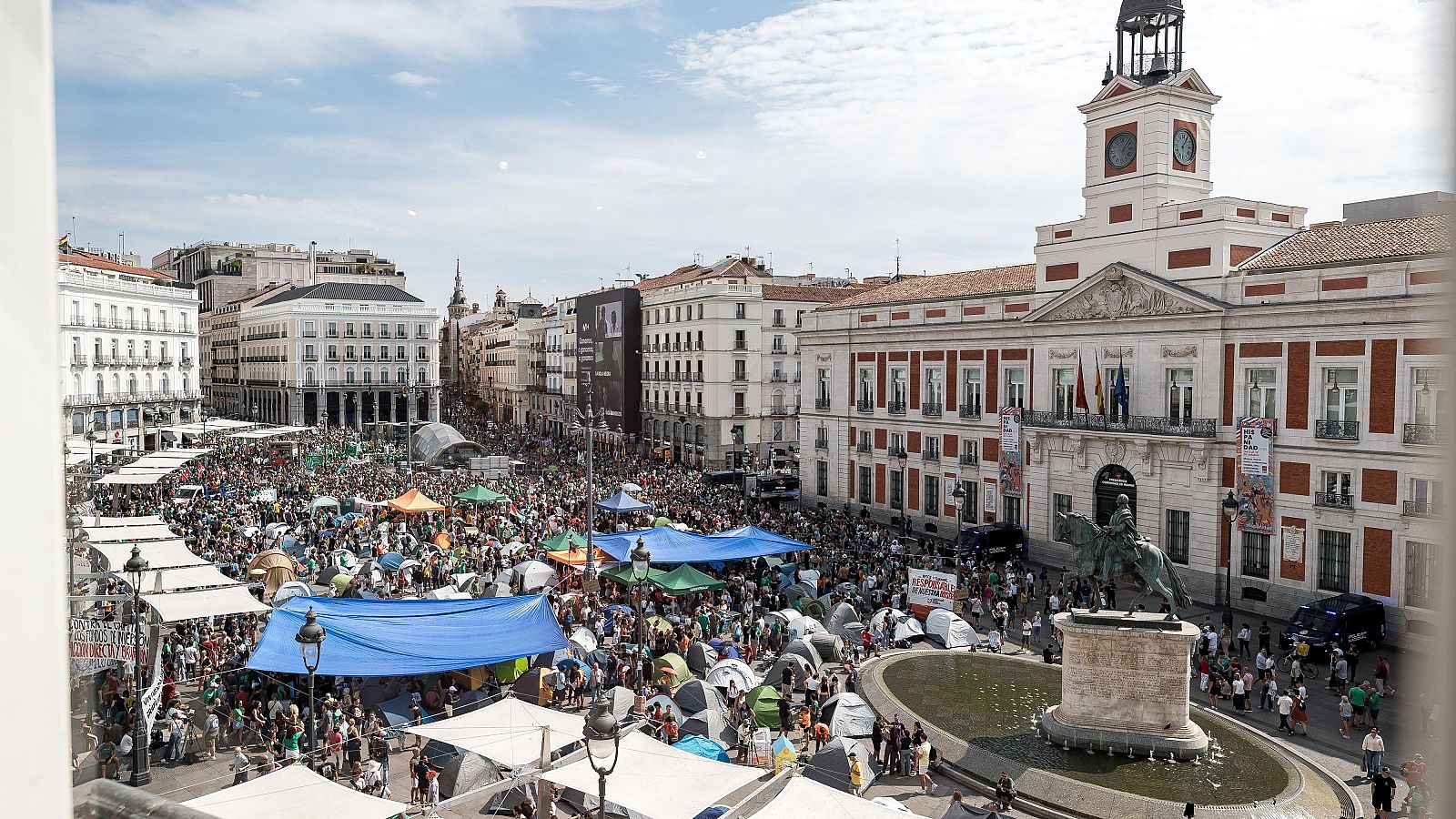 Vista aérea de la Puerta del Sol abarrotada de gente, con tiendas de campaña y lonas, y la Real Casa de Correos al fondo.