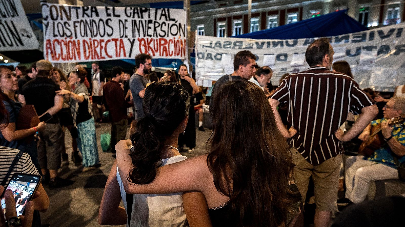 Multitud en protesta nocturna urbana con pancartas "ACCIÓN DIRECTA Y OCUPACIÓN" y "UNIVERSIDAD PÚBLICA". Un hombre toca un instrumento de cuerda mientras otros conversan y se apoyan mutuamente.
