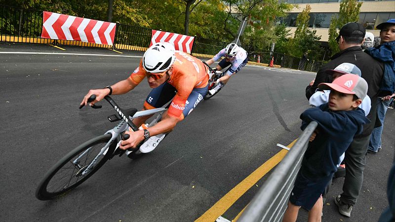Ciclistas en plena curva de una carrera. Uno viste naranja y azul con "Rabobank", el otro sigue de cerca. Se ven espectadores y barreras de seguridad al fondo.