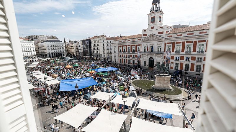 Vista aérea de la Puerta del Sol con una masiva acampada y multitud. Se distinguen tiendas, lonas, la Real Casa de Correos y la estatua de Carlos III.