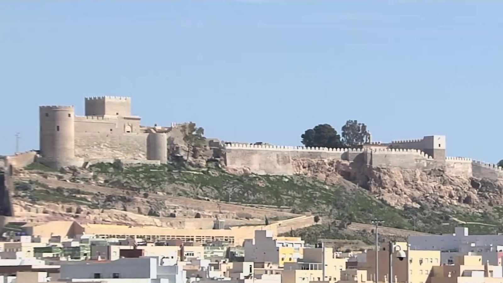 Vista panorámica de la Alcazaba de Almería, una fortaleza histórica sobre una colina rocosa, con murallas, torres y una ciudad a sus pies bajo un cielo azul.