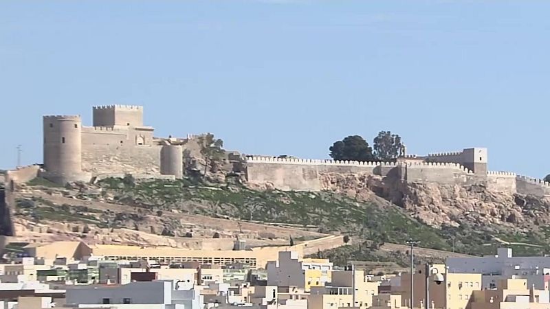 Vista panorámica de la Alcazaba de Almería, una fortaleza histórica sobre una colina rocosa, con murallas, torres y una ciudad a sus pies bajo un cielo azul.