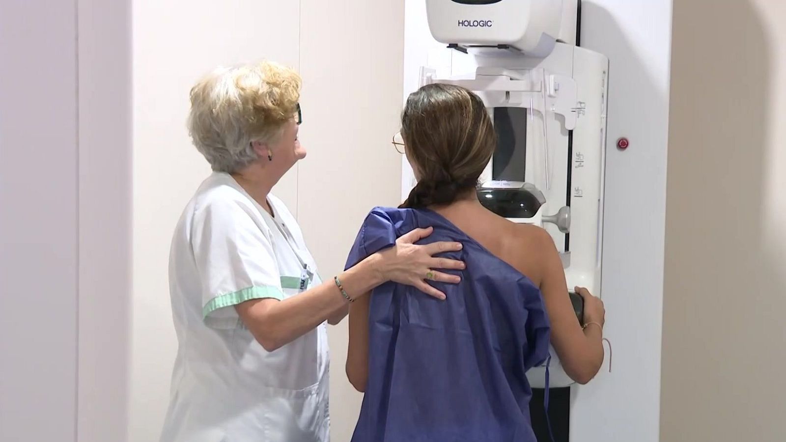 Profesional sanitaria y paciente en un examen médico. La máquina "HOLOGIC" es visible. La sanitaria apoya su mano en la espalda de la paciente.