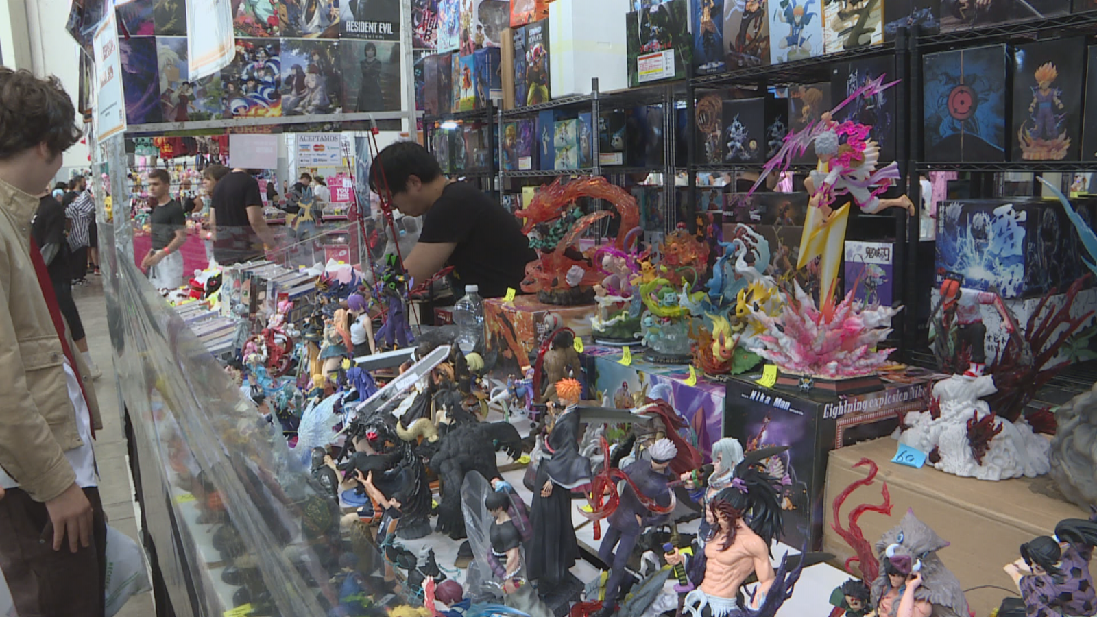 Festival de manga y cultura pop con un expositor repleto de figuras de anime y manga. Se aprecian personajes como Ichigo Kurosaki y Satoru Gojo, además de un póster de "RESIDENT EVIL".