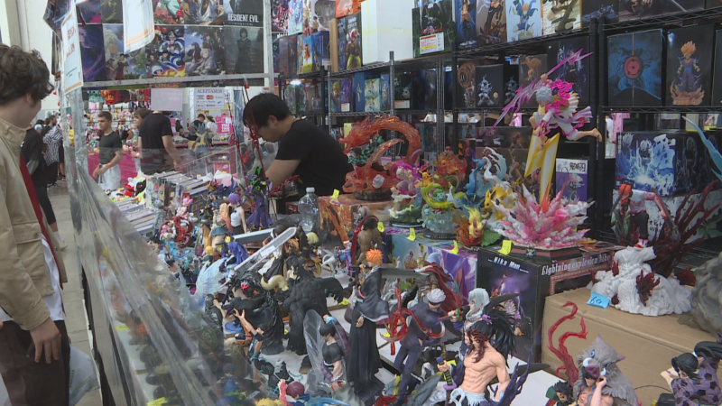 Festival de manga y cultura pop con un expositor repleto de figuras de anime y manga. Se aprecian personajes como Ichigo Kurosaki y Satoru Gojo, además de un póster de "RESIDENT EVIL".