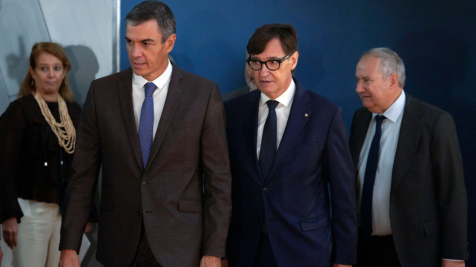 Pedro Sánchez, Salvador Illa y Jordi Hereu, figuras políticas españolas, caminan en un evento. Sánchez viste traje marrón grisáceo, Illa gafas y traje azul oscuro, Hereu traje oscuro y camisa azul claro.