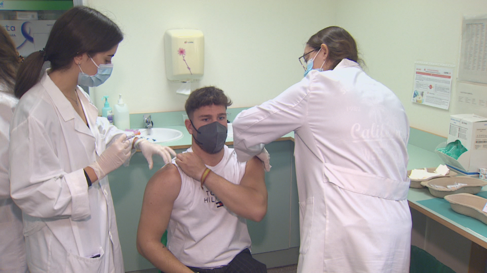 Profesionales sanitarias vacunan a un joven con camiseta "TOMMY HILFIGER" en un centro médico. Se aprecian jeringuilla, batas, mascarillas y suministros médicos.