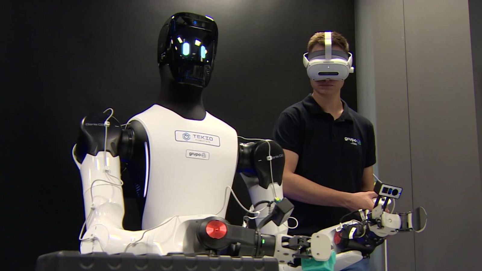 Robot humanoide con logotipos "TEKIO" y "grupo i" junto a un hombre con visor VR y controlador, sugiriendo teleoperación en un entorno oscuro.